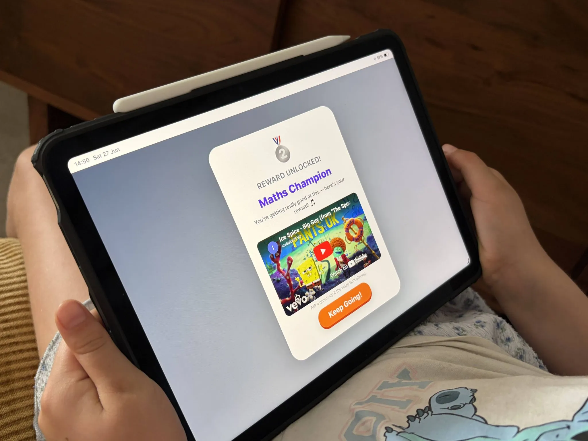 A child holding an iPad showing a reward unlocked screen reading Maths Champion, above a music video she chose as the reward.